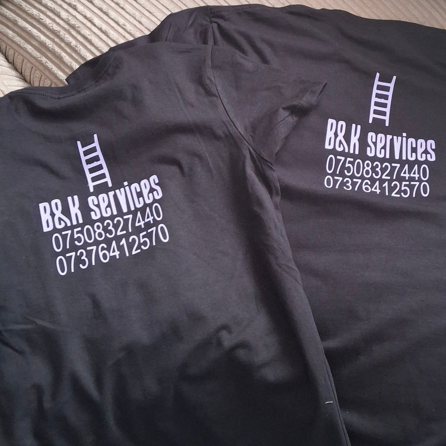 Two black t-shirts laid out on a ribbed beige sofa, both printed with white text reading 'B&K SERVICES' and a phone number.