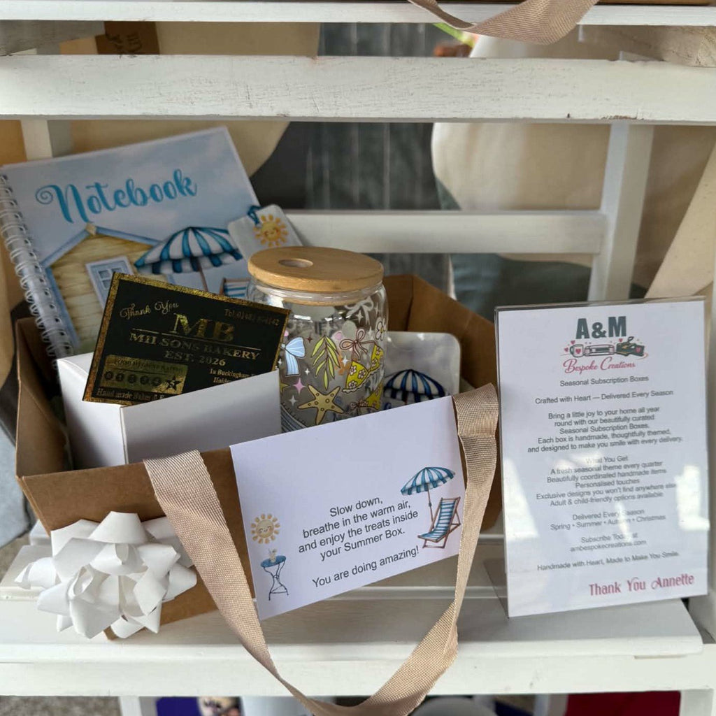 A gift basket arrangement on a white shelf containing a notebook, a Milson Bakery Small Nibble Box of brownies, a small card, a glass libby with a summer design exclusive to the subscription boxes, a printed thank-you card, and a personalised coaster and matching bookmark