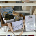 A gift basket arrangement on a white shelf containing a notebook, a Milson Bakery Small Nibble Box of brownies, a small card, a glass libby with a summer design exclusive to the subscription boxes, a printed thank-you card, and a personalised coaster and matching bookmark