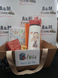 A cardboard gift basket filled with school-themed items, including a white insulated water bottle with a cartoon girl illustration, a notebook, a chocolate treat box, and other small accessories. The basket has a beige handle and a white sign reading 'You've Got This' with school icons.