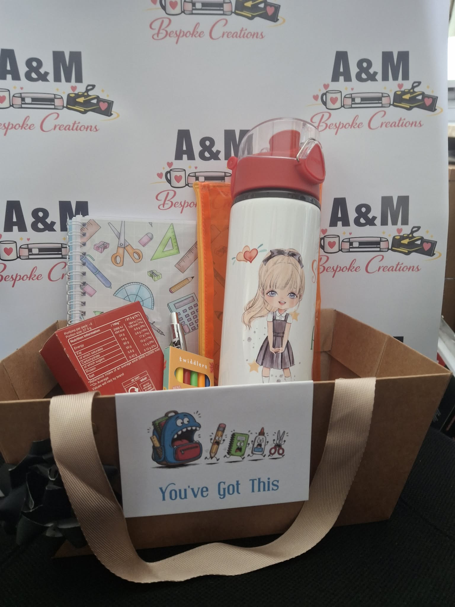 A cardboard gift basket filled with school-themed items, including a white insulated water bottle with a cartoon girl illustration, a notebook, a chocolate treat box, and other small accessories. The basket has a beige handle and a white sign reading 'You've Got This' with school icons.