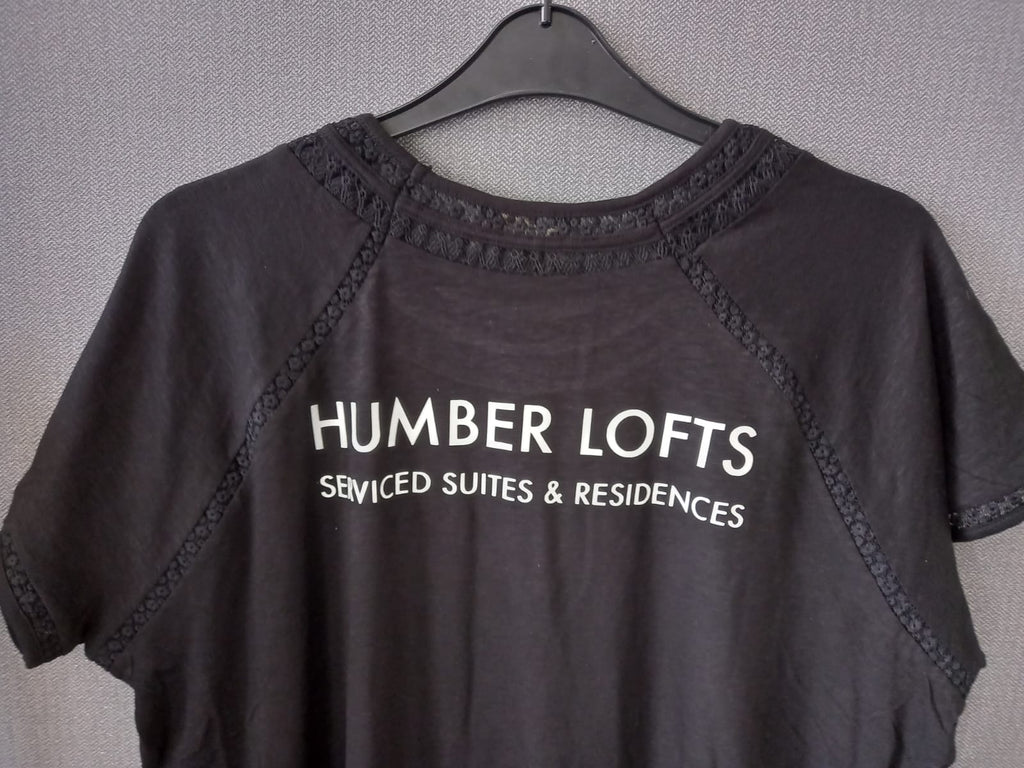A dark charcoal gray short-sleeve t-shirt hanging on a black hanger against a textured gray wall. The shirt features white printed text reading 'HUMBER LOFTS SERVICED SUITES & RESIDENCES' across the chest, with decorative lace trim along the neckline and raglan seams.