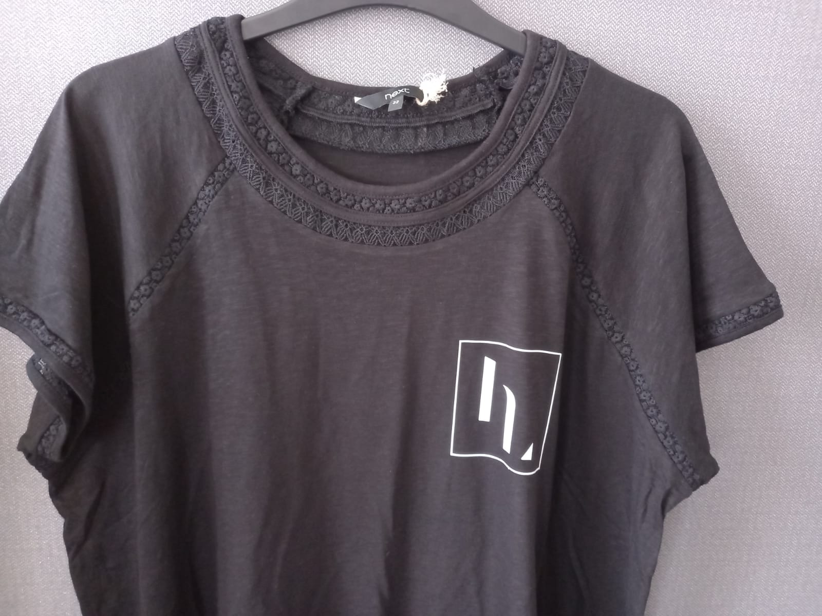 A dark gray short-sleeve raglan t-shirt with lace trim around the crew neck and sleeves, featuring a white graphic logo on the left chest, hanging on a gray hanger against a light gray textured wall.