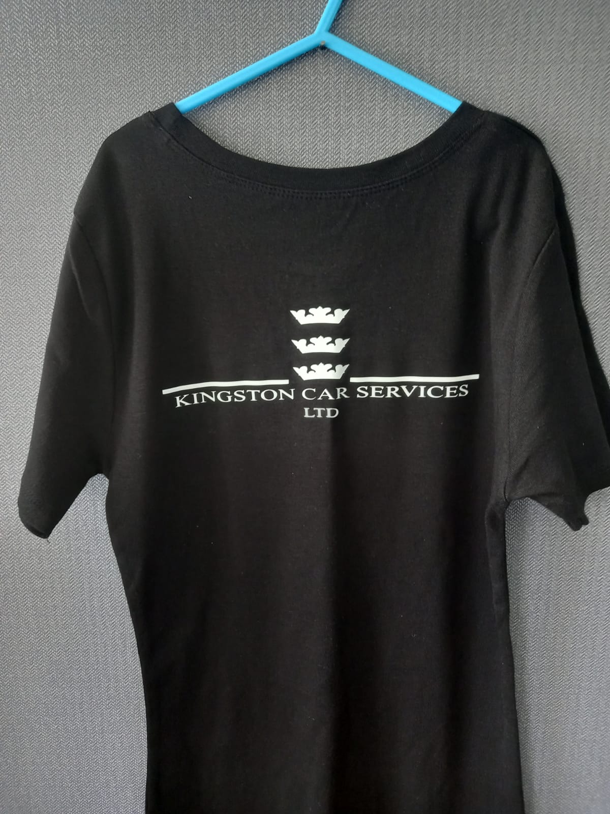 A black short-sleeve t-shirt hanging on a bright blue hanger against a gray textured wall. The shirt features a white printed graphic on the chest with three crowns, a horizontal line, and the text 'KINGSTON CAR SERVICES LTD'.