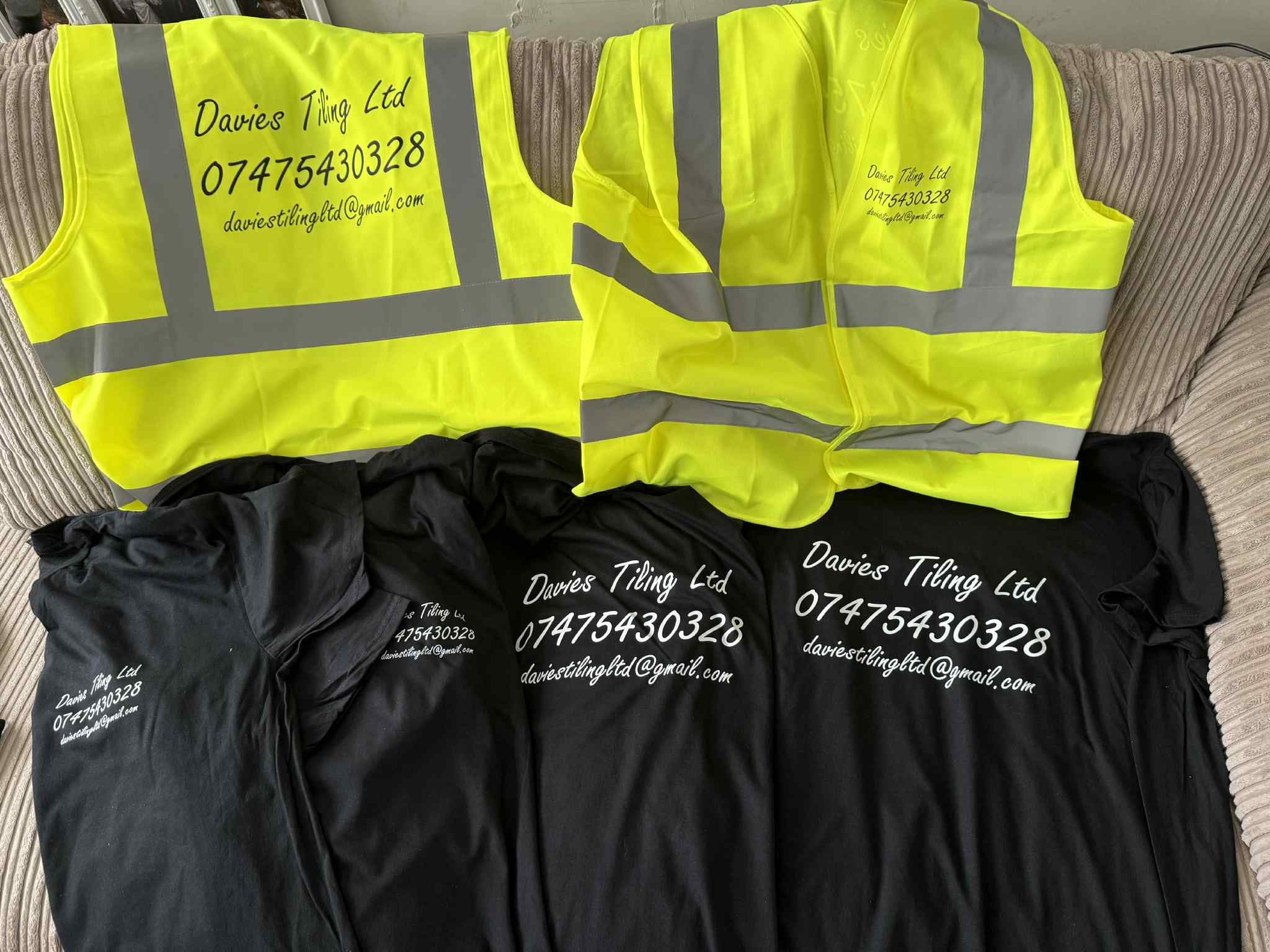 Two high-visibility neon yellow jackets with reflective gray stripes, laid flat on a striped fabric surface, with three black t-shirts behind them showing white printed text.