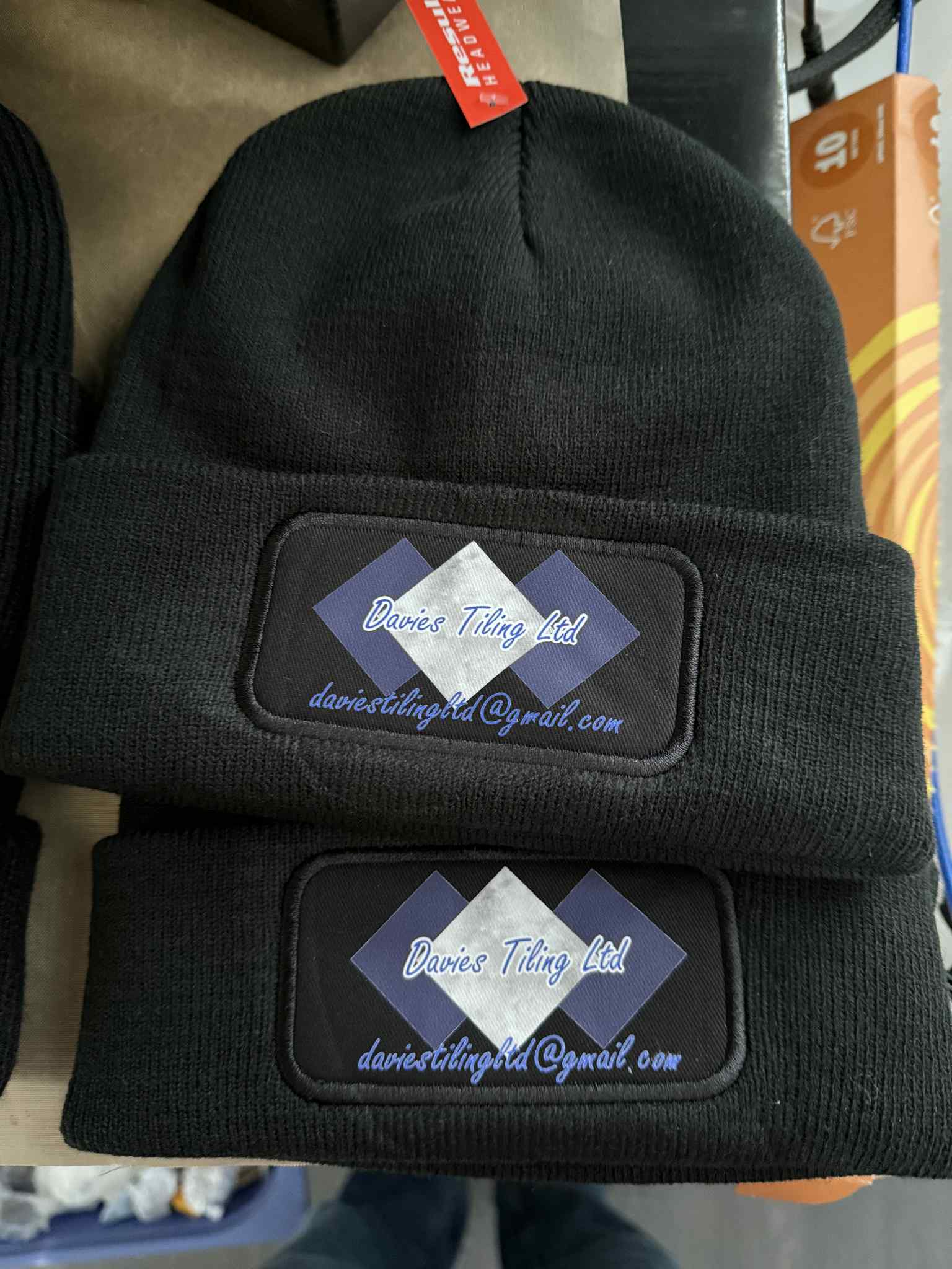Two black beanie hats stacked together, each with a rectangular embroidered patch on the cuff. The patch has a diamond pattern in blue, white, and black, with the text 'Bureau Thing Ltd' and 'daniesthing99@gmail.
