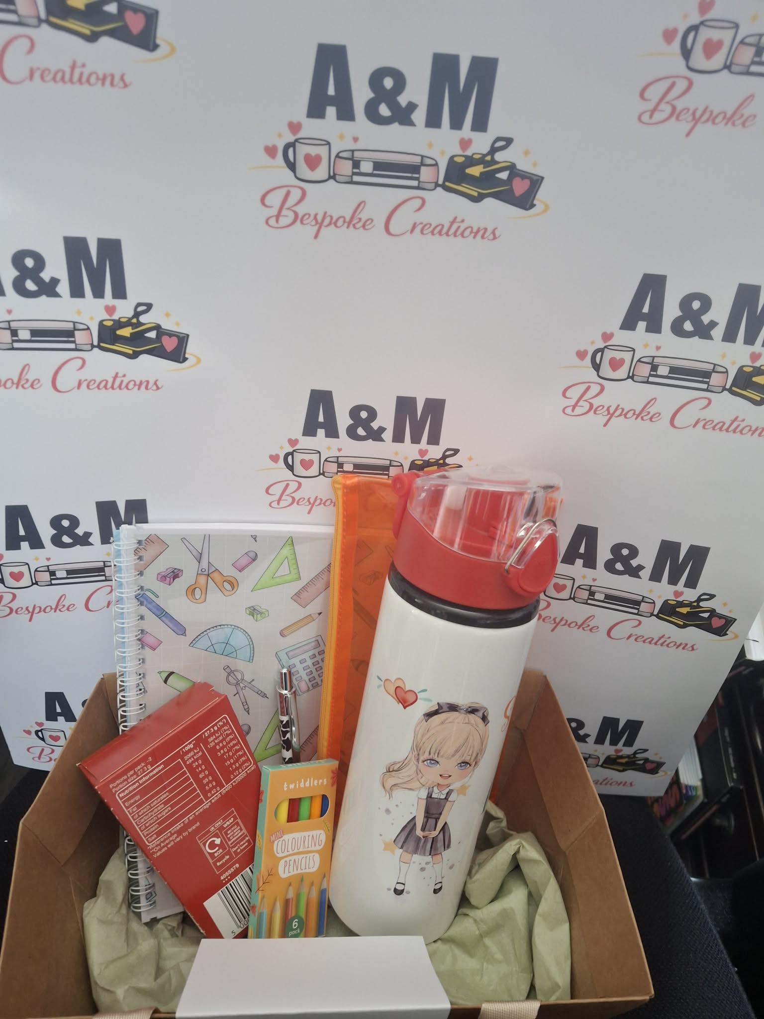A curated gift box filled with stationery and accessories, including a white insulated bottle with a red lid, a notebook, pencils, and a treat box, set against a branded A&M Bespoke Creations backdrop.