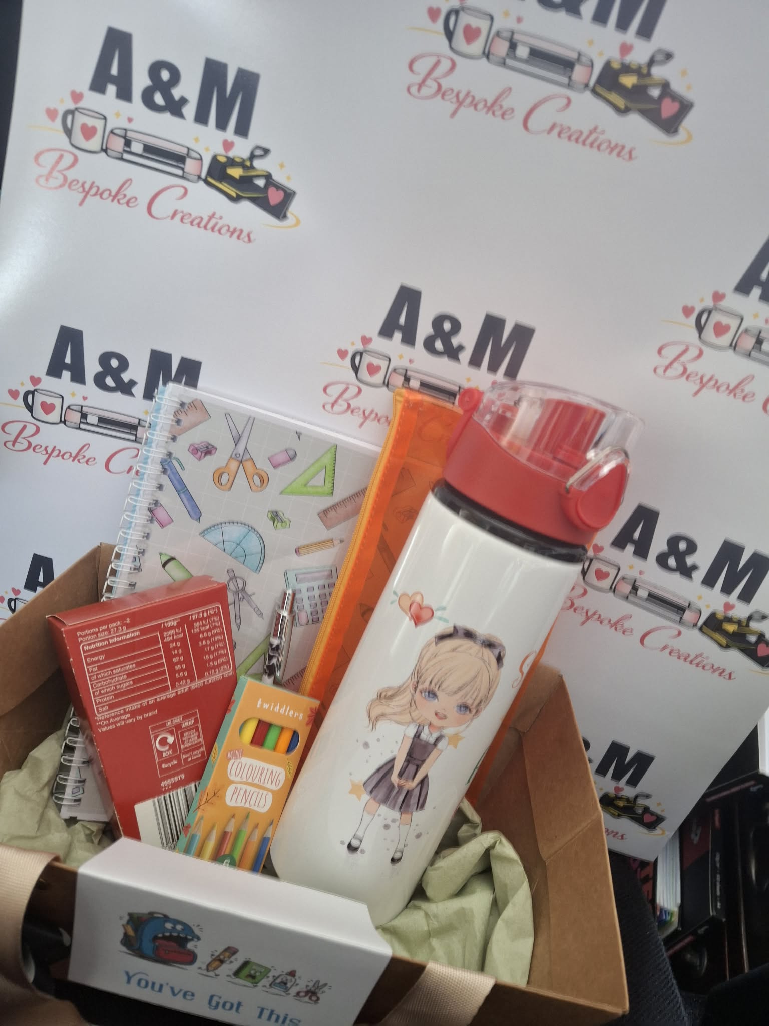 A curated gift box containing a white water bottle with a red lid and anime character print, a notebook, a treat box, and other stationery items, set against a branded A&M Bespoke Creations backdrop.
